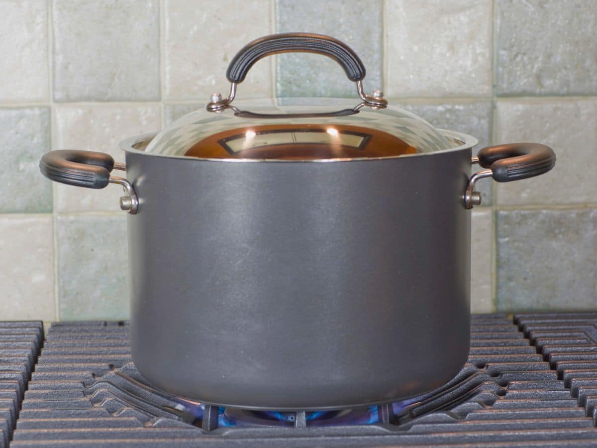 Large black pot with a lid on a gas stove, displaying visible blue flames, against a tiled backsplash.