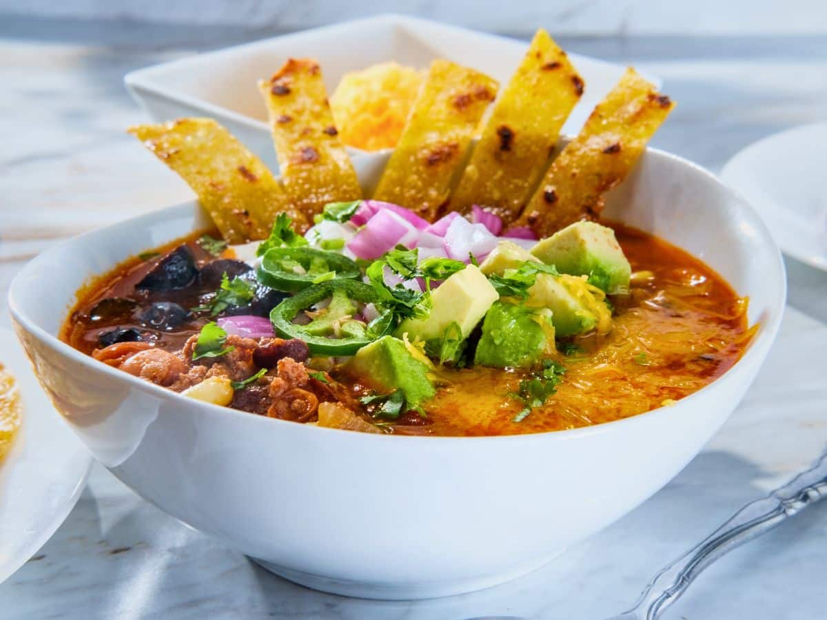 A bowl of spicy soup topped with diced avocado, onions, jalapeños, and crispy tortilla strips on a marble surface.