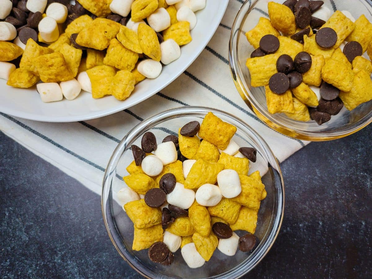 Three bowls containing a snack mix of cereal squares, mini marshmallows, and chocolate chips, placed on a striped cloth.