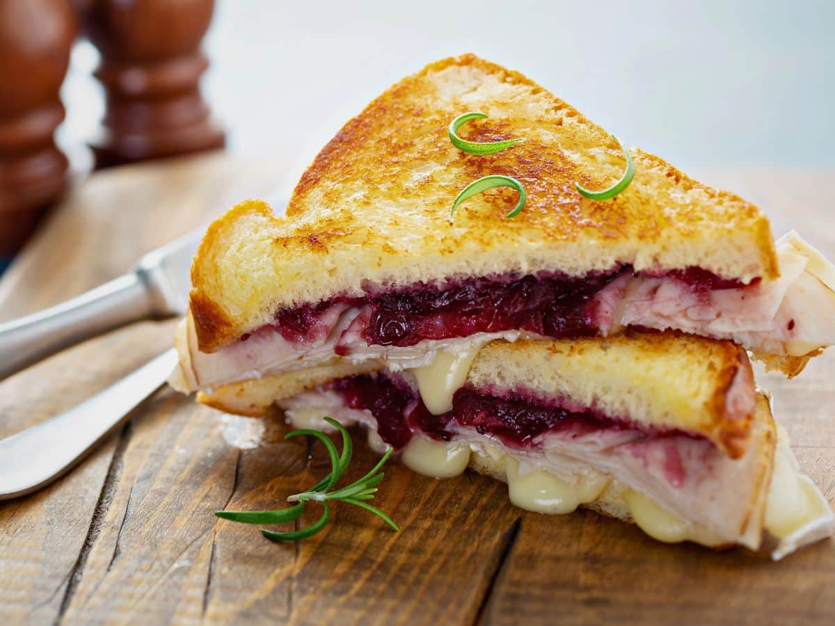 Grilled cheese sandwich with melted cheese, turkey, and cranberry sauce, garnished with rosemary on a wooden surface.
