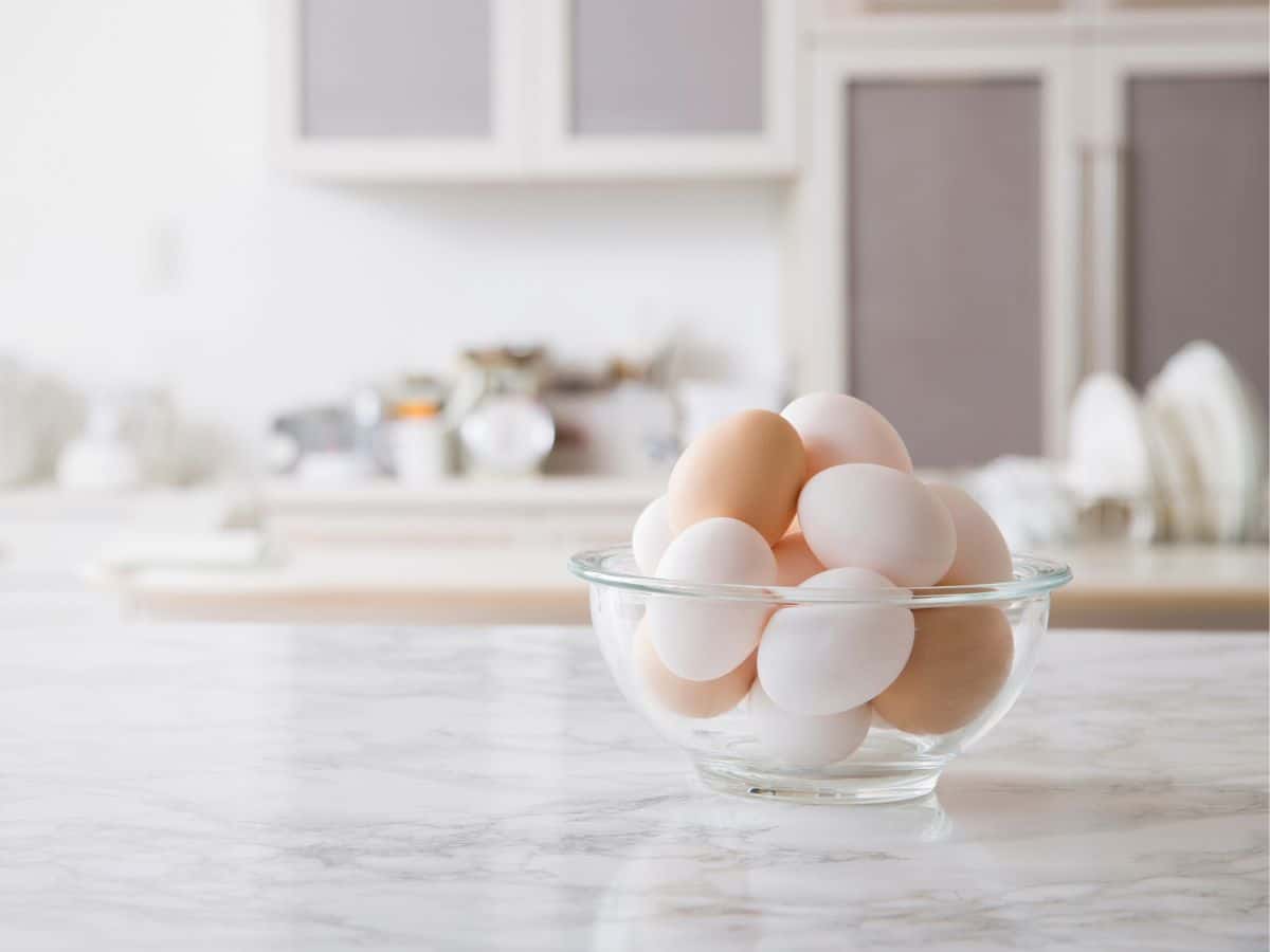 A glass bowl filled with white and brown eggs sits on a marble countertop in a kitchen.