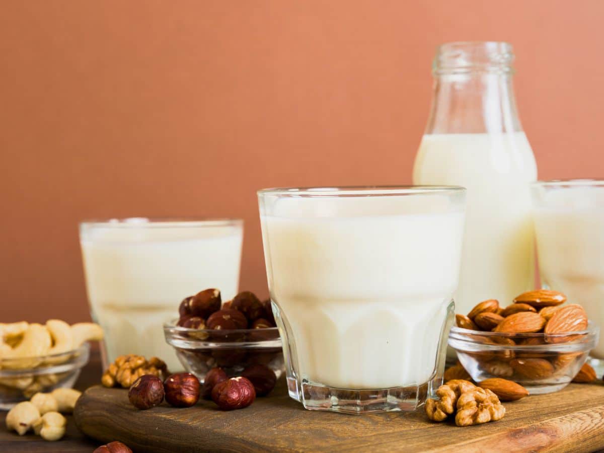 Glasses and a bottle of plant-based milk on a wooden board, surrounded by nuts including almonds, walnuts, and hazelnuts. Brownish background.