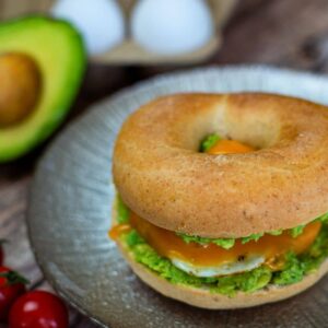Bagel with avocado, sunny-side-up egg, and cheese on a plate. Avocado half, eggs, and cherry tomatoes in the background.