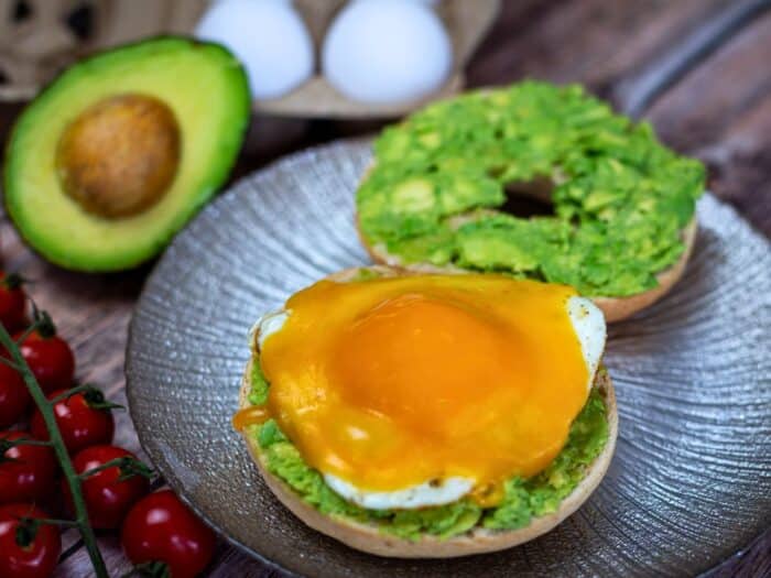 A bagel topped with mashed avocado, a fried egg, and melted cheese on a plate. A whole avocado, eggs, and cherry tomatoes are in the background.