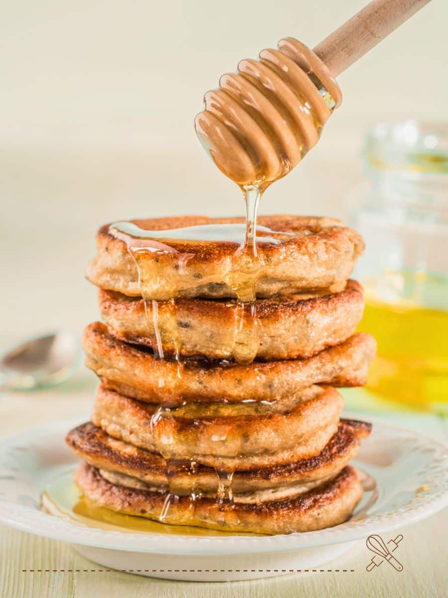 A stack of pancakes with honey being drizzled from a honey dipper, set on a white plate. A jar of honey and a spoon are in the background.