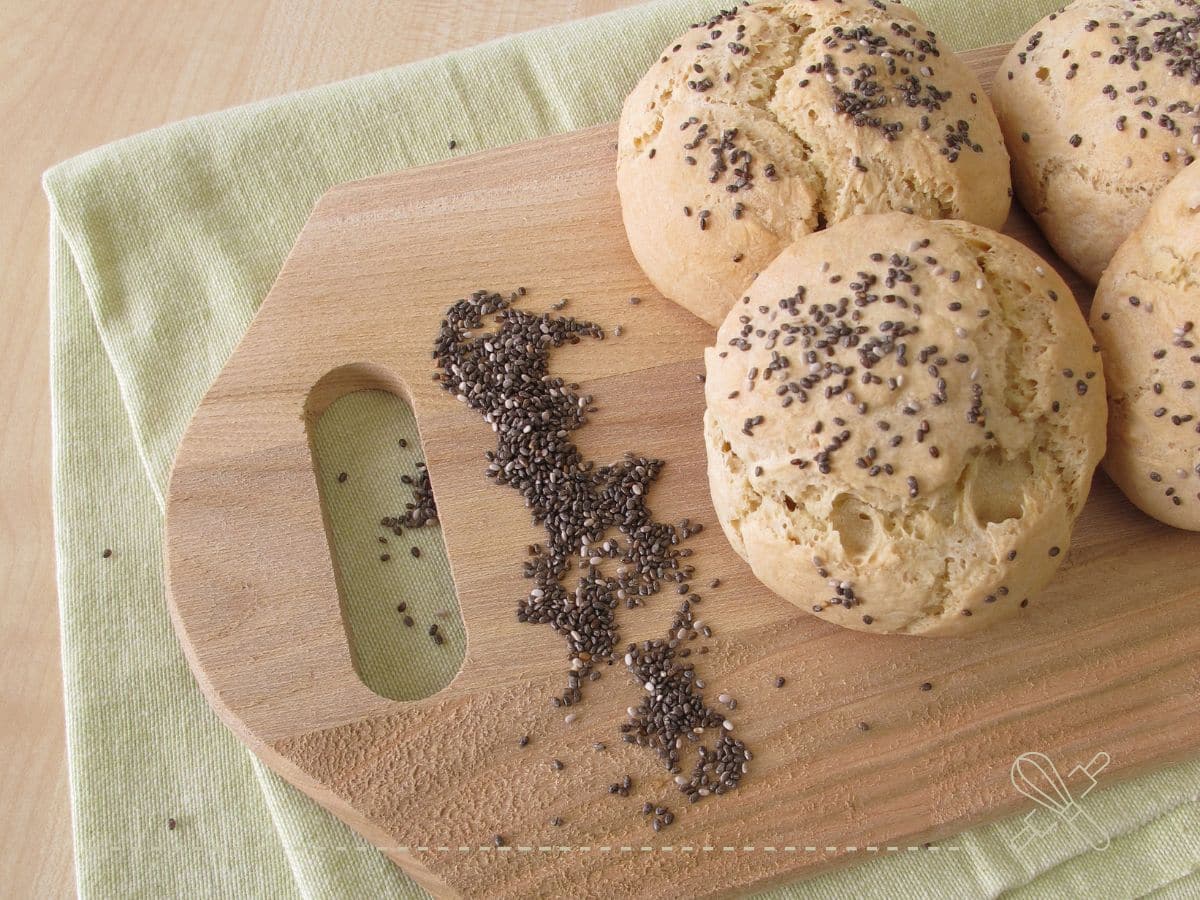 Four chia seed-topped buns are placed on a wooden cutting board, with some seeds scattered on the board.