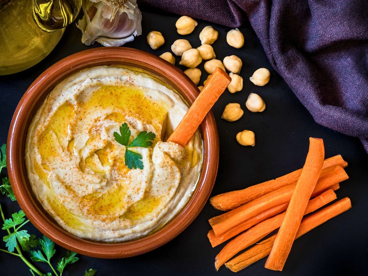 A bowl of hummus with olive oil and a parsley leaf, surrounded by chickpeas, garlic, and carrot sticks on a dark surface.