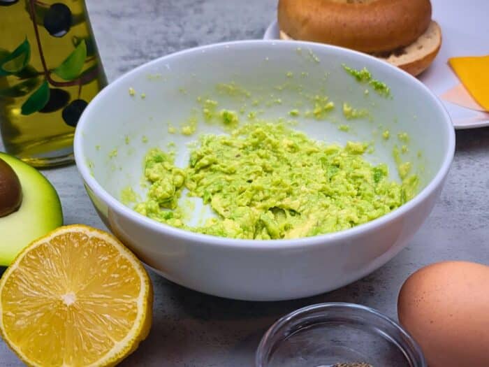 A white bowl of mashed avocado next to a lemon half, egg, avocado half, and a bagel on a table.
