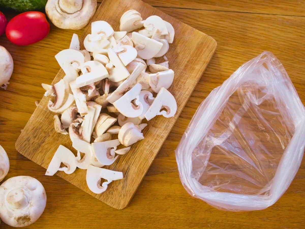 Sliced mushrooms on a wooden cutting board beside an empty plastic bag on a wooden surface.