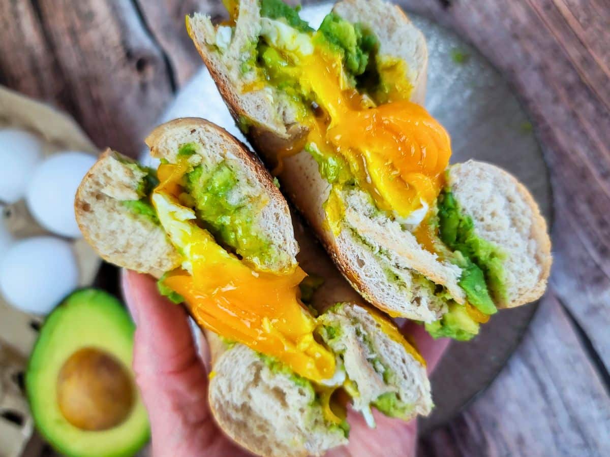 A split bagel with avocado and oozing egg yolk held above a wooden surface, with an avocado half and eggs nearby.