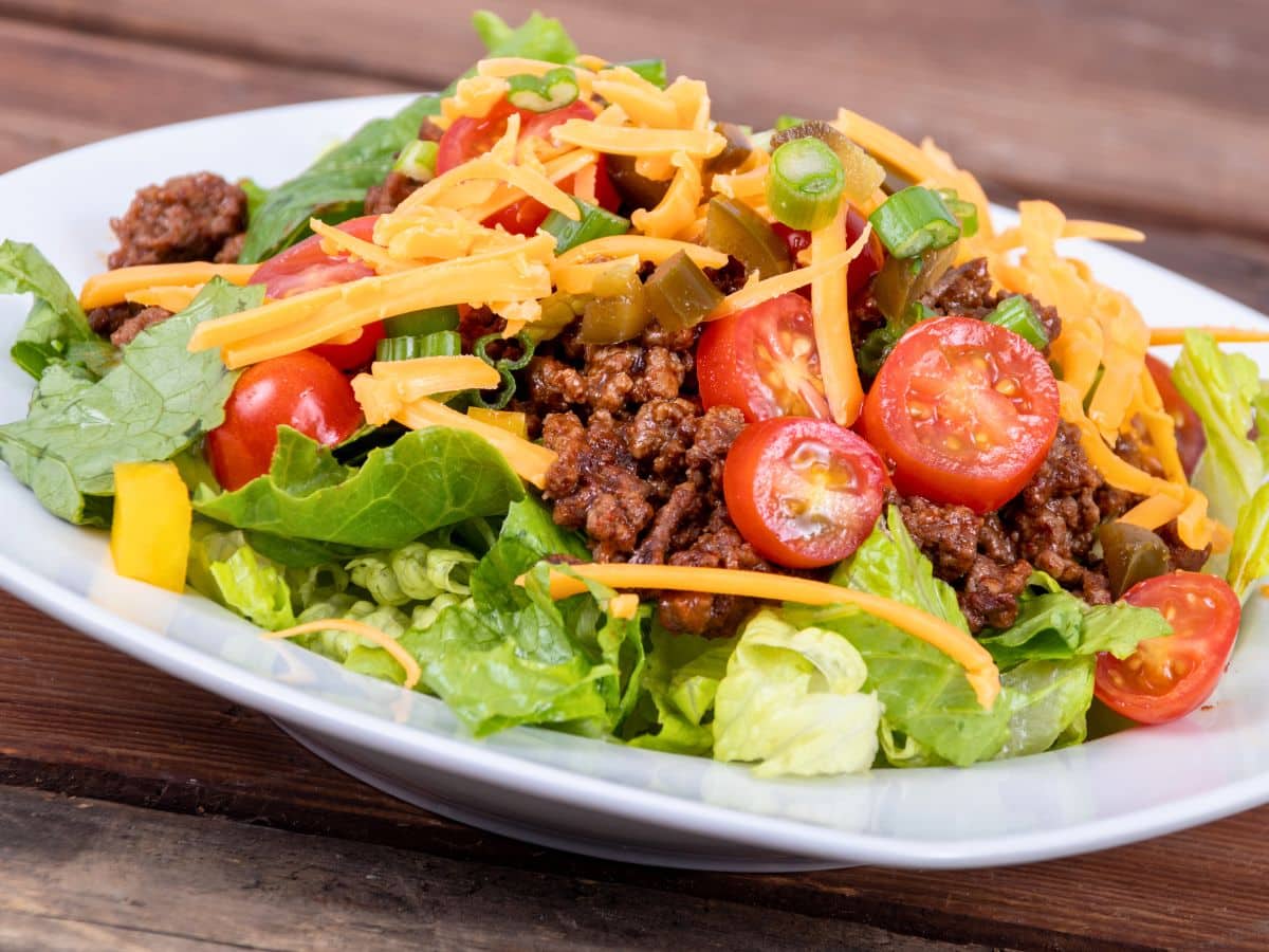A white plate with a taco salad featuring ground meat, shredded cheddar cheese, cherry tomatoes, lettuce, yellow bell peppers, and green onions.