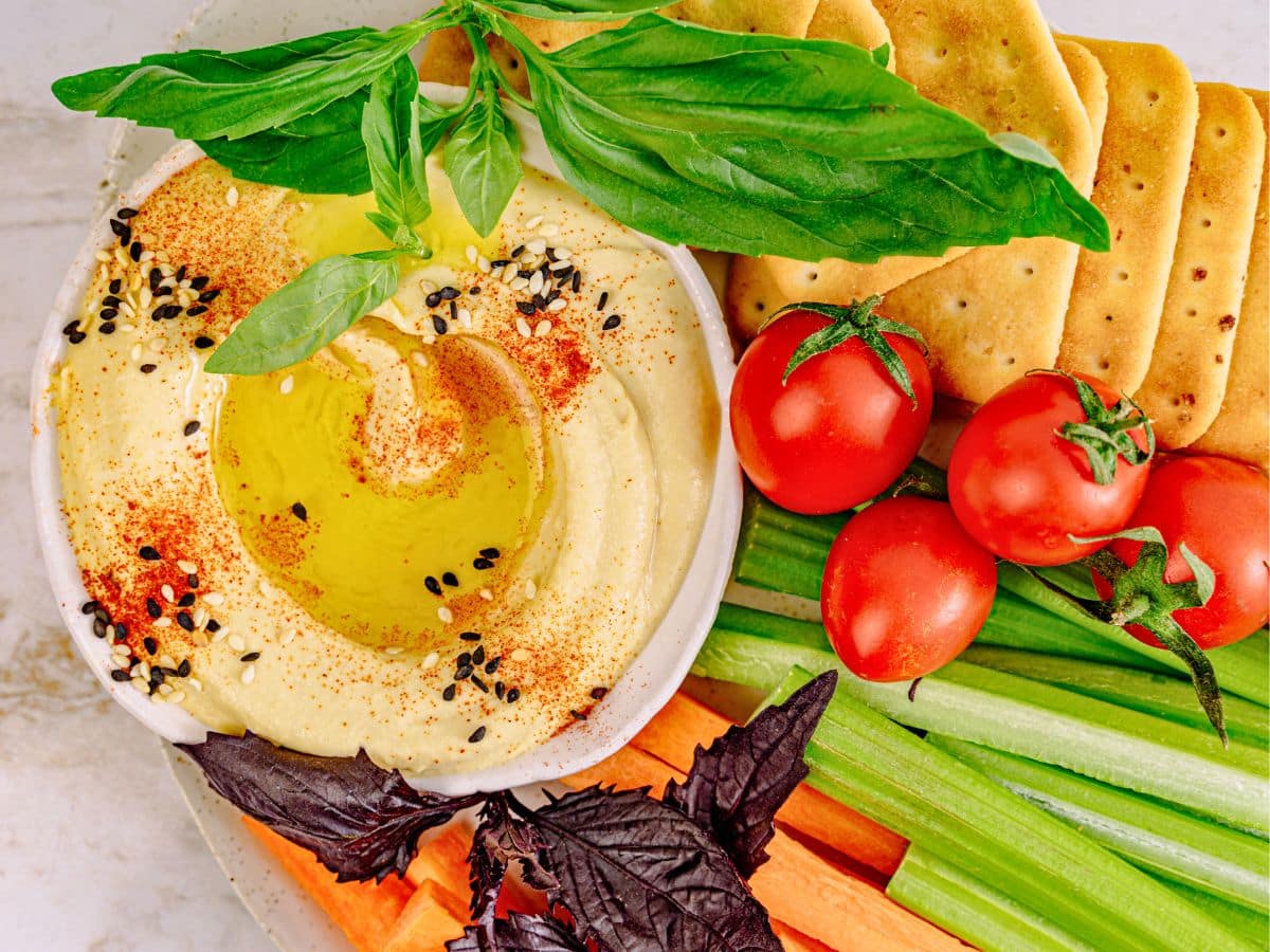 A bowl of hummus with olive oil and spices, surrounded by basil, cherry tomatoes, crackers, carrot, and celery sticks.
