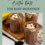 Three jars of chocolate peanut butter oats topped with cream and chocolate chips, placed on a wooden board with a spoon and scattered peanuts and chips nearby.