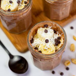Glass jars with chocolate pudding topped with sliced bananas, shredded coconut, and chocolate chips on a wooden board, with a black spoon and scattered peanuts nearby.