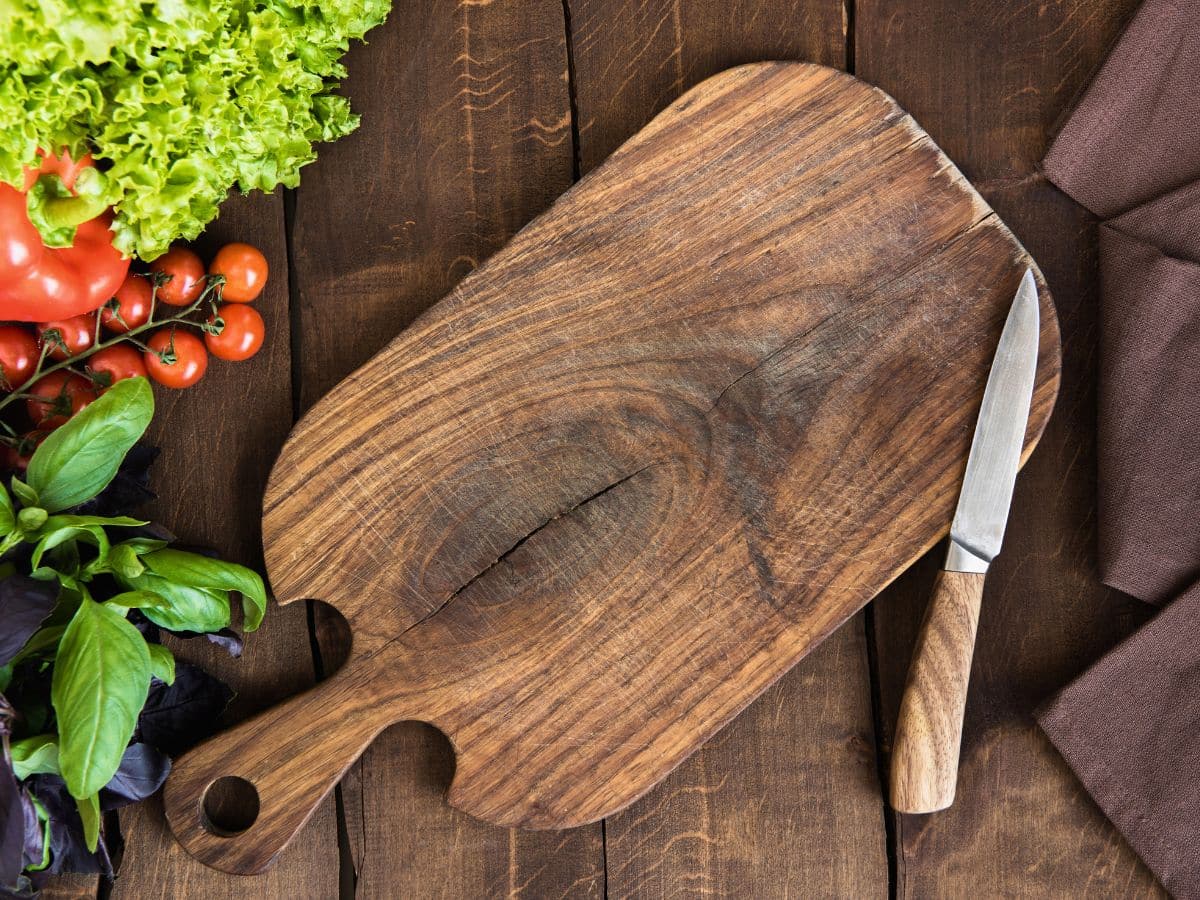 Wooden cutting board with a knife on a dark wooden table, surrounded by lettuce, cherry tomatoes, basil, and a brown cloth napkin.
