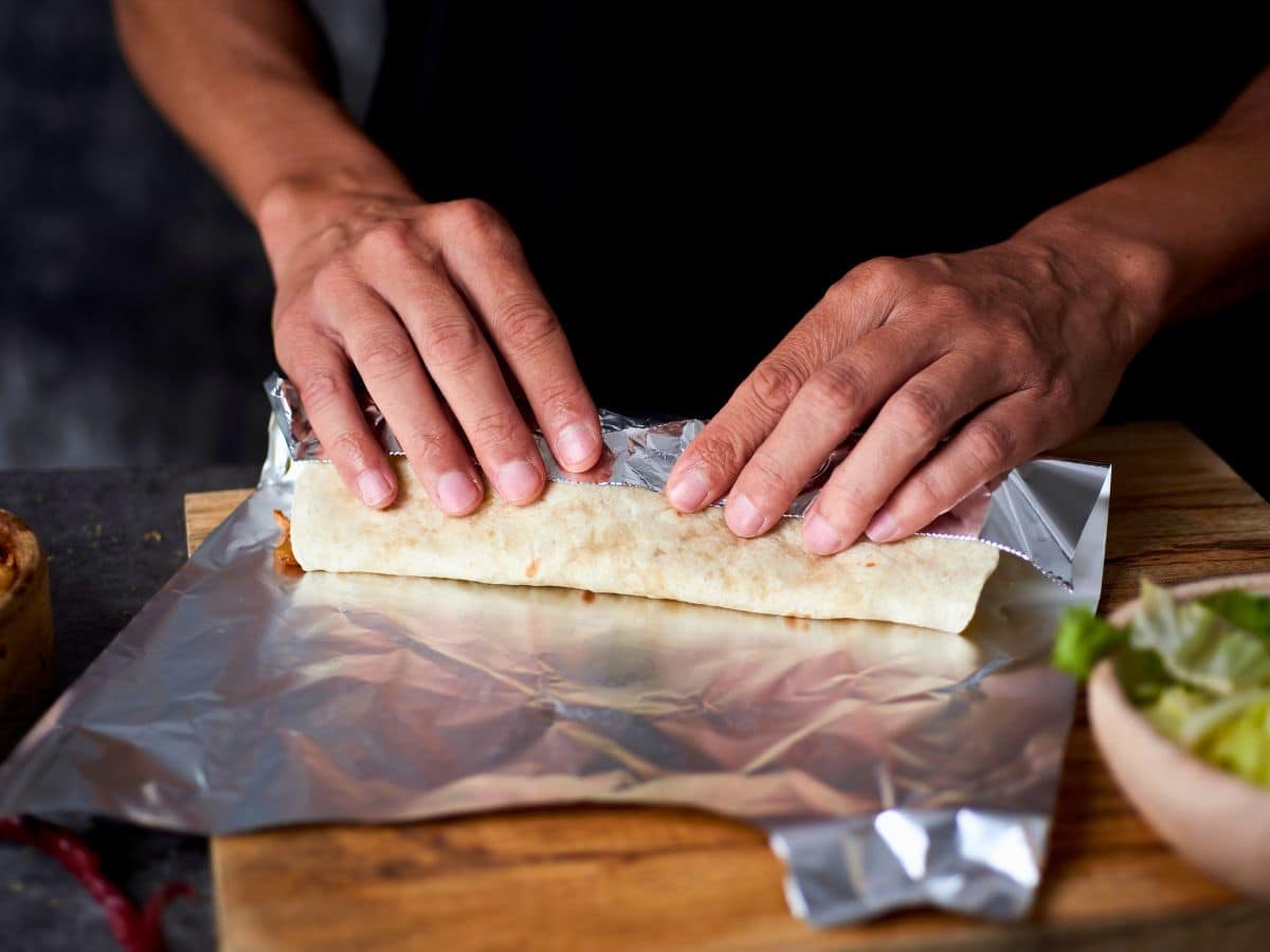 Hands wrapping a burrito in aluminum foil on a wooden board.