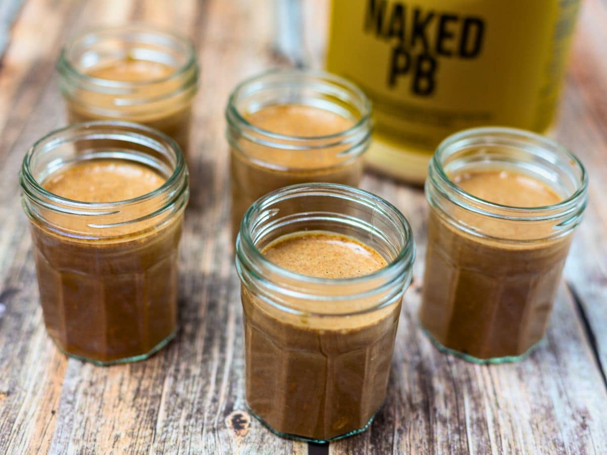 Six small glass jars filled with a brown liquid sit on a wooden surface, with a container labeled "Naked PB" visible in the background.