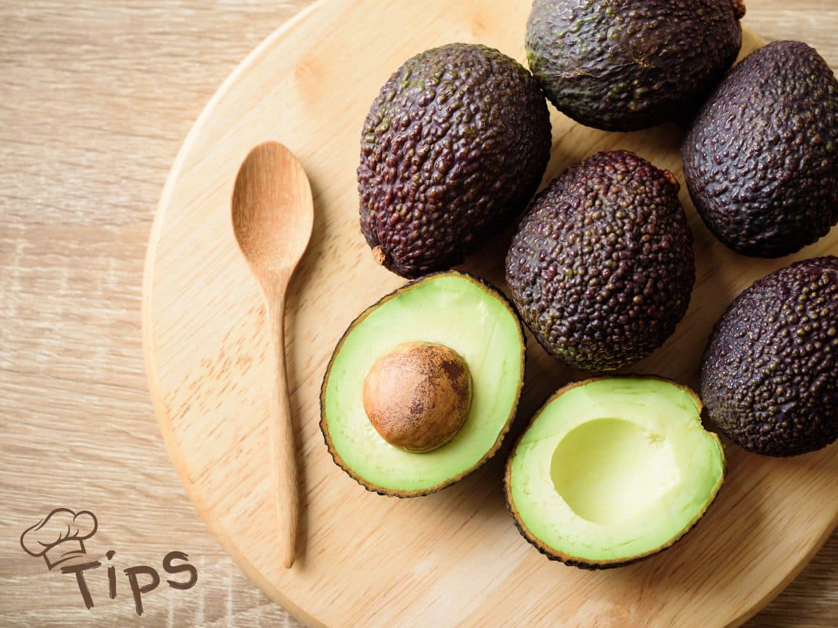 Several ripe avocados on a round wooden board, one split open showing the seed. A wooden spoon is placed beside them. The word "Tips" is visible on the lower left.