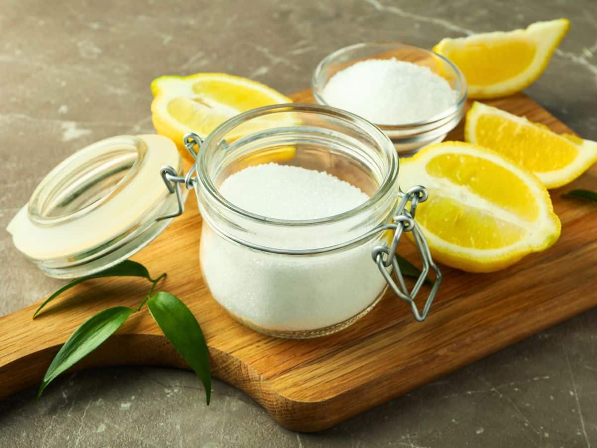 Glass jar and bowls of white sugar on a wooden board with lemon slices and green leaves.