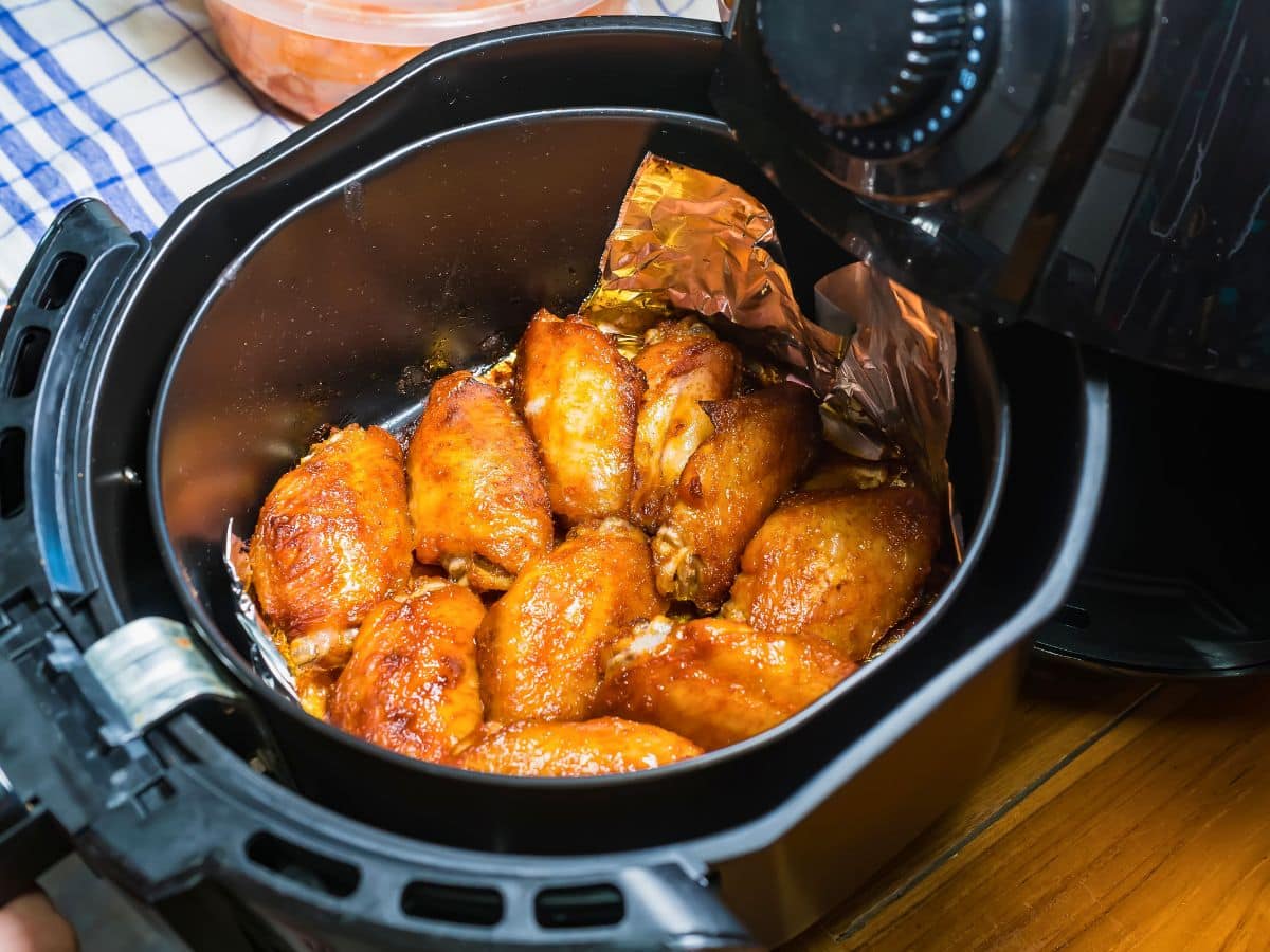 Chicken wings in an air fryer, partially lined with foil, on a wooden surface.