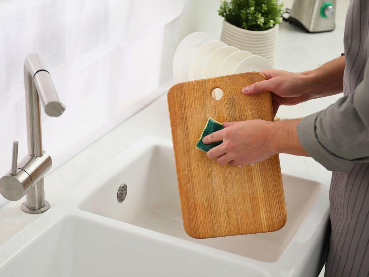 Person cleaning a wooden cutting board over a kitchen sink with a green sponge.