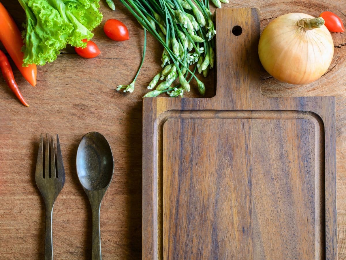 Wooden cutting board on a table with lettuce, chili peppers, cherry tomatoes, spring onions, a whole onion, and wooden fork and spoon nearby.