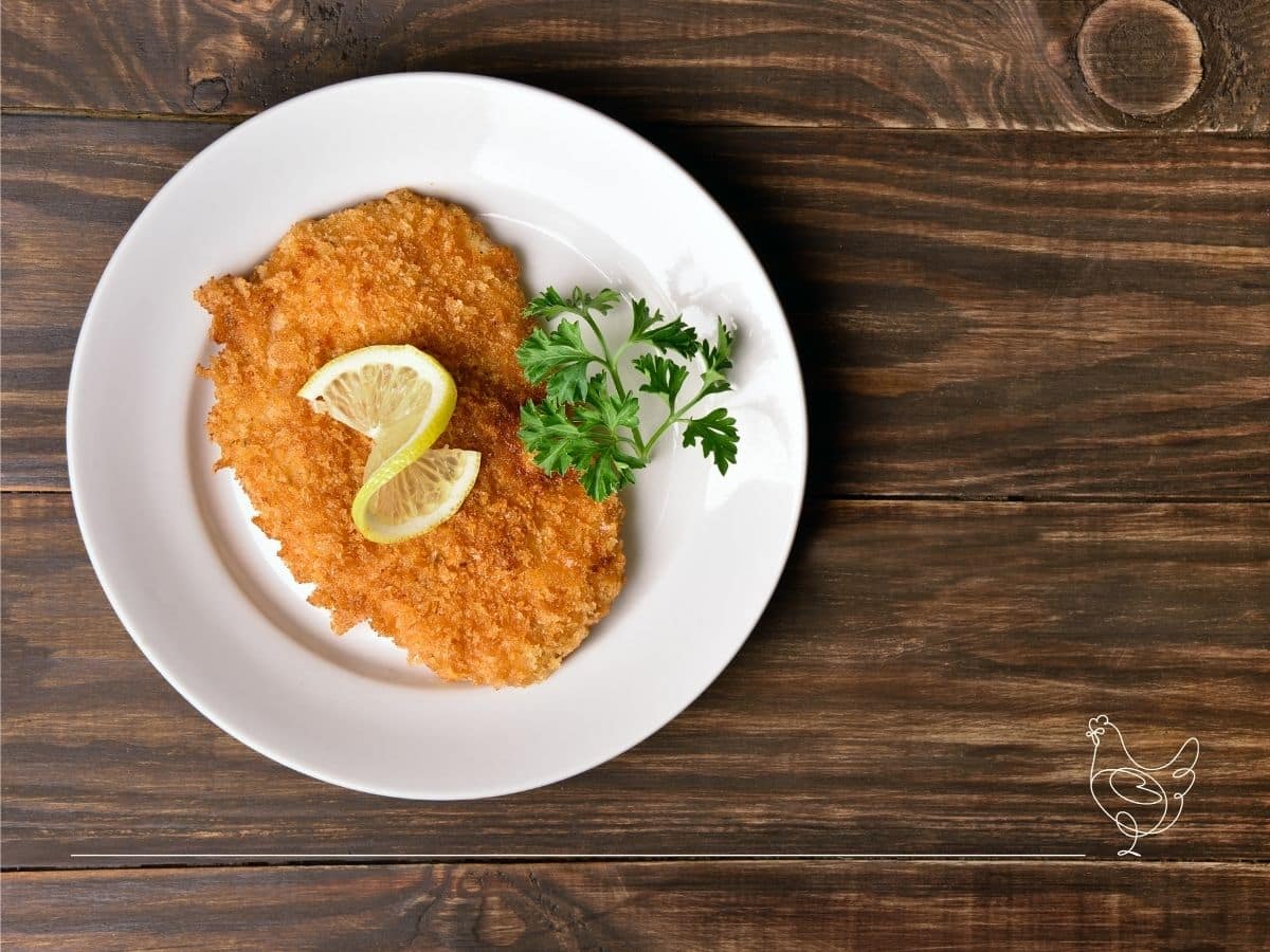 A breaded and fried cutlet on a white plate, garnished with two lemon wedges and parsley, placed on a wooden table.