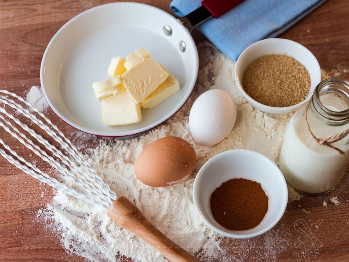 Ingredients for baking on a wooden surface: butter, eggs, sugar, milk, flour, cinnamon, a whisk, and a spatula.