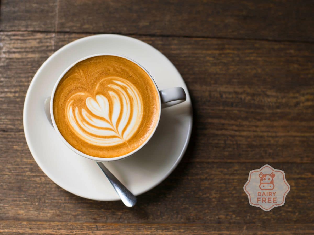 A cup of latte with heart-shaped latte art on a saucer, placed on a wooden table. A "Dairy Free" label is visible near the cup.