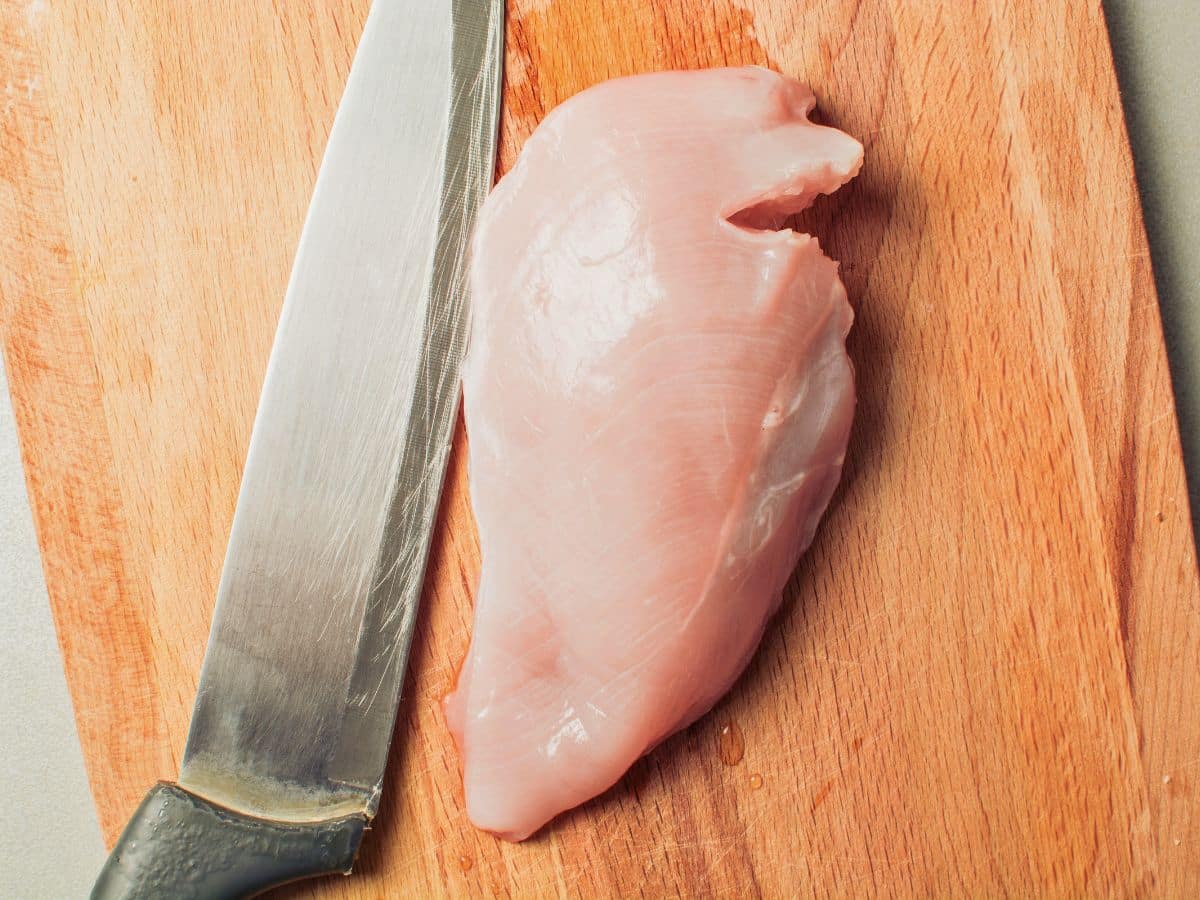 Raw chicken breast and a large kitchen knife on a wooden cutting board.