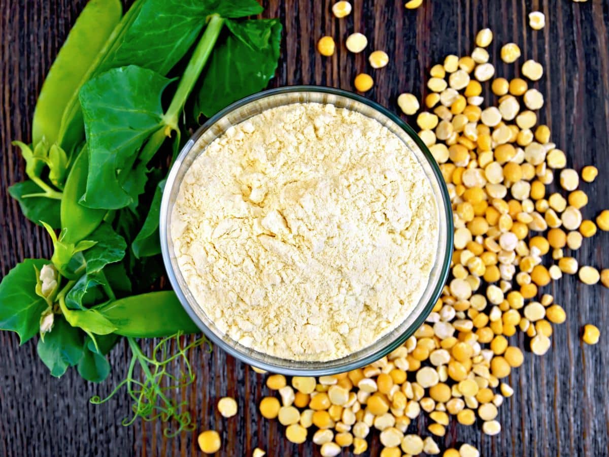 A bowl of yellow pea flour is surrounded by whole dried peas and green pea pods on a wooden surface.