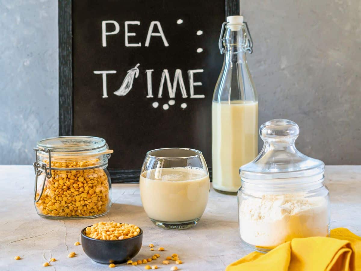 Chalkboard with "PEA TIME," bottle of milk, glass of milk, jar of split peas, small bowl of peas, and a jar of flour on a table with a yellow napkin.