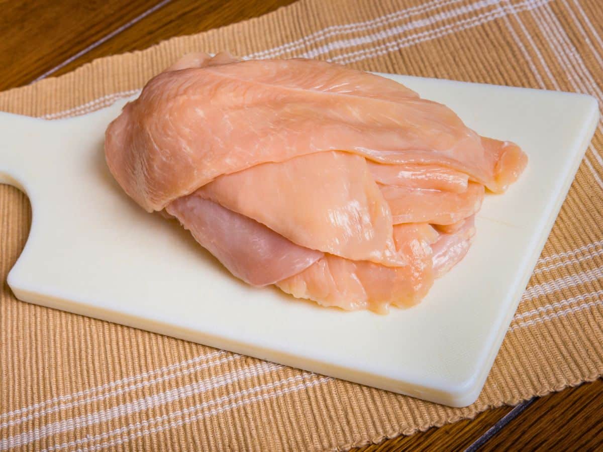Raw chicken breast pieces on a white cutting board, placed on a tan cloth with striped details.