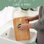 Person cleaning a wooden cutting board with a sponge over a sink. Text reads, "Clean Your Wooden Cutting Board Like a Pro.