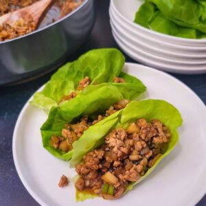 Three lettuce wraps filled with a ground meat and vegetable mixture are served on a white plate, with extra lettuce leaves and filling in the background.