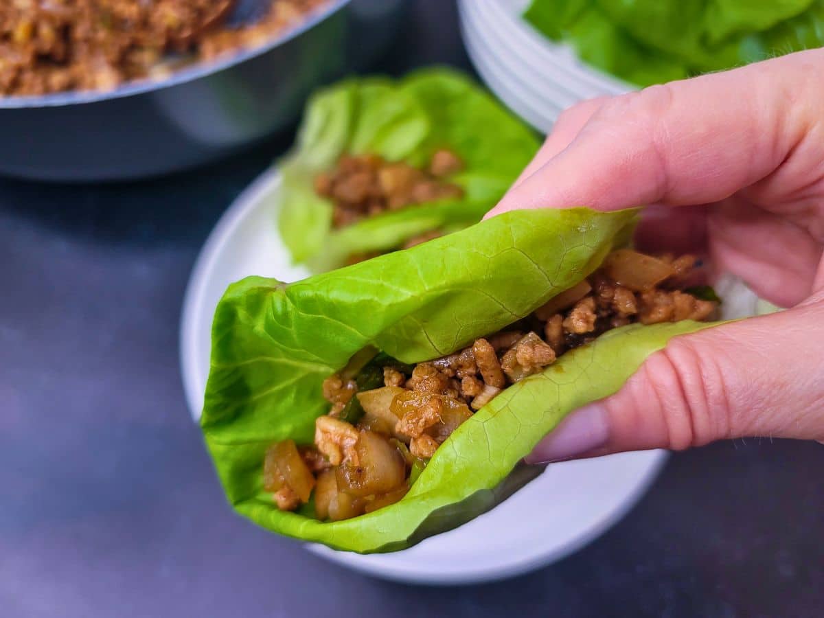 A hand holds a lettuce wrap filled with seasoned ground meat and diced vegetables, with more lettuce wraps and a pan in the background.