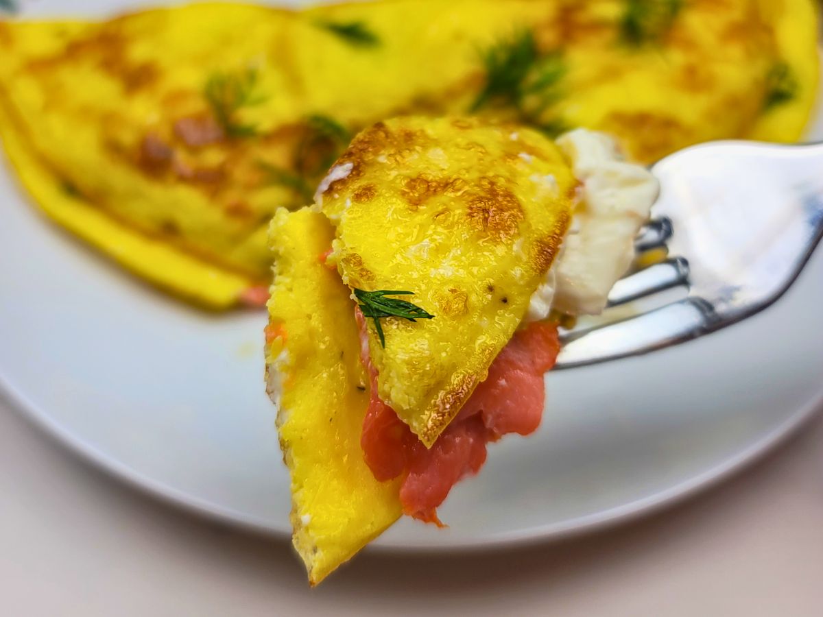 A close-up of a fork holding a piece of omelette with visible salmon, cream cheese, and dill, with the rest of the omelette on a white plate in the background.