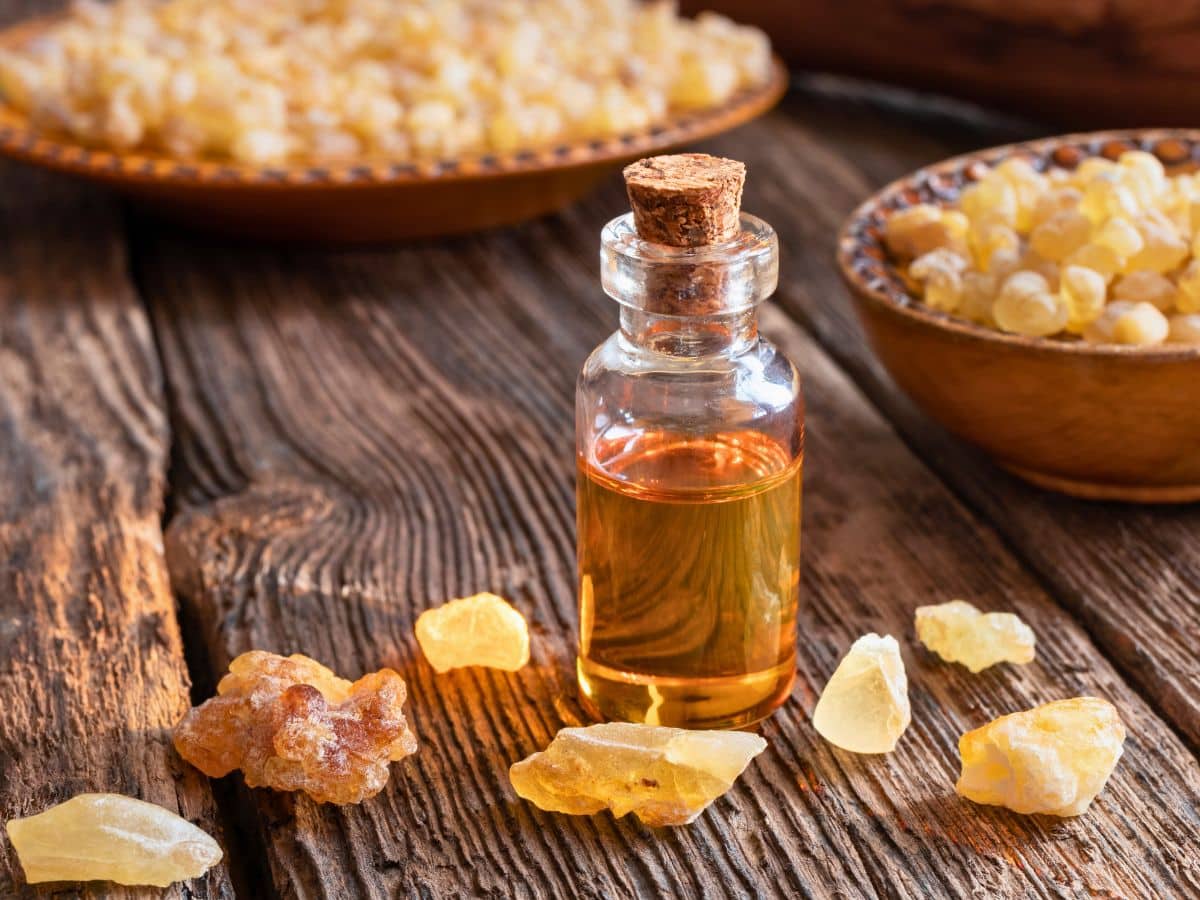 A small glass bottle of amber-colored oil with a cork stopper sits on a wooden surface, surrounded by chunks of yellow and white resin. Bowls of resin are visible in the background.
