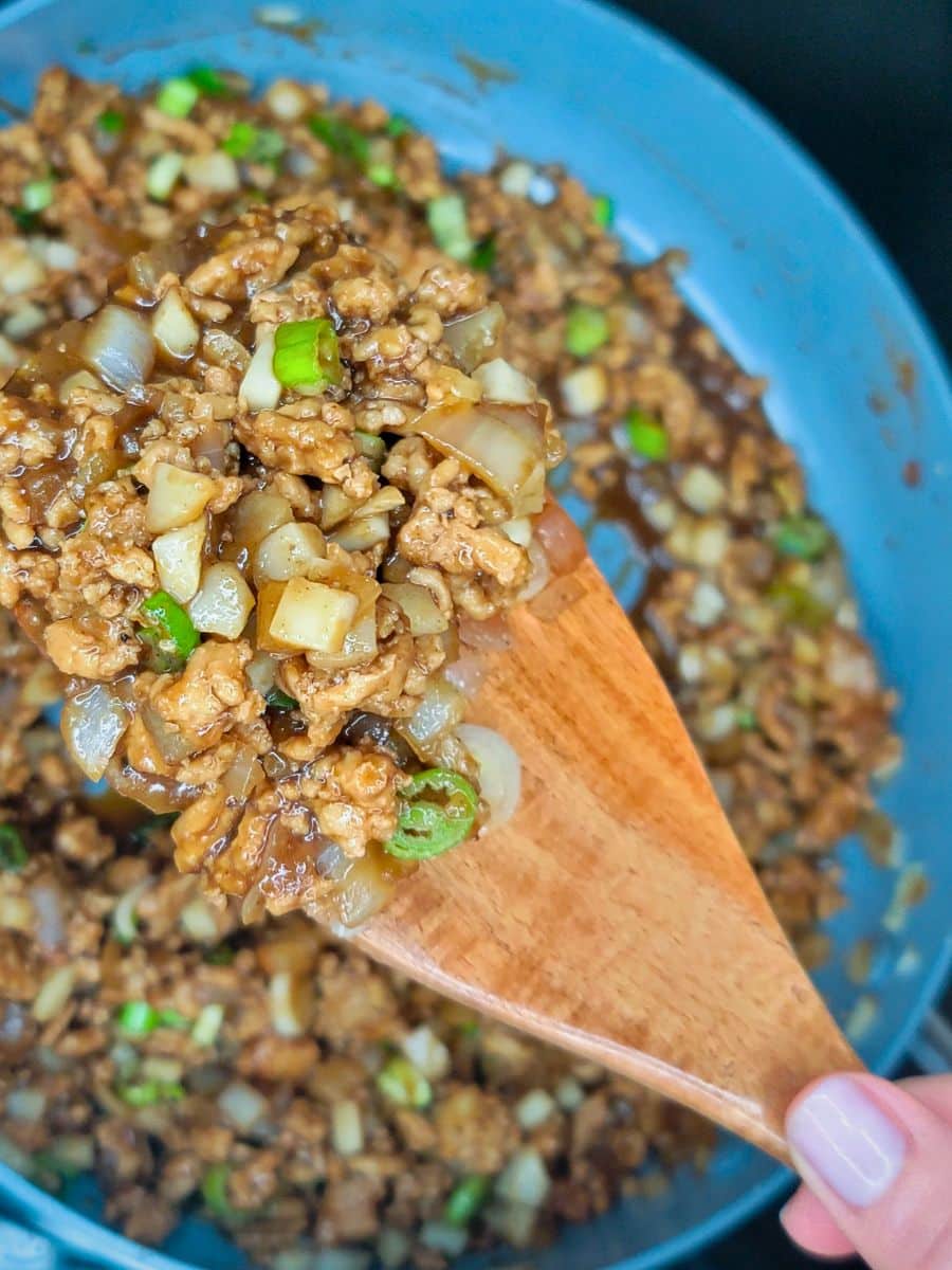 A wooden spoon holds a mixture of cooked ground meat, chopped onions, and green onions over a pan containing the same mixture.