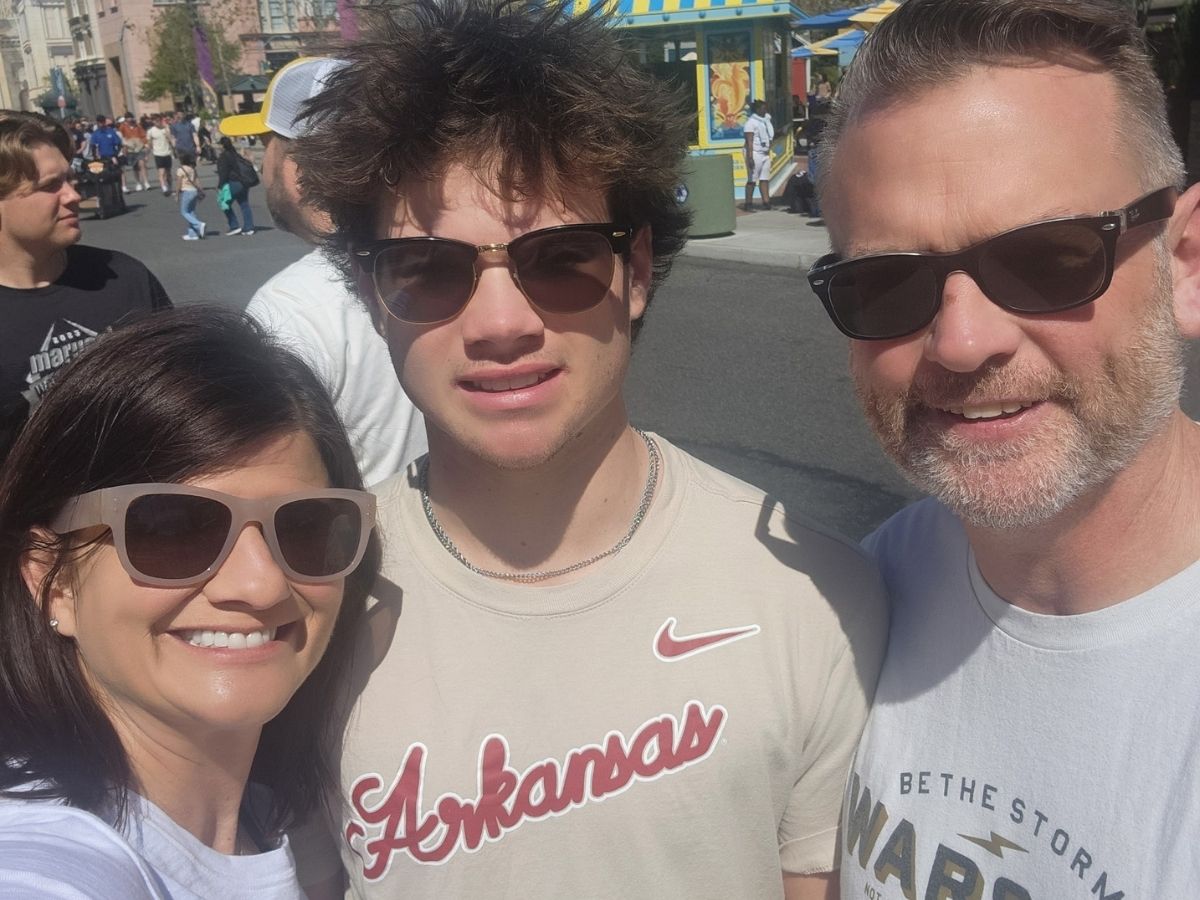 Three people wearing sunglasses pose for a selfie outdoors on a sunny day. The person in the middle wears an "Arkansas" shirt. There are other people and colorful buildings in the background.
