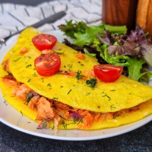 A folded omelette filled with salmon and herbs, topped with halved cherry tomatoes, served with mixed greens on a white plate.