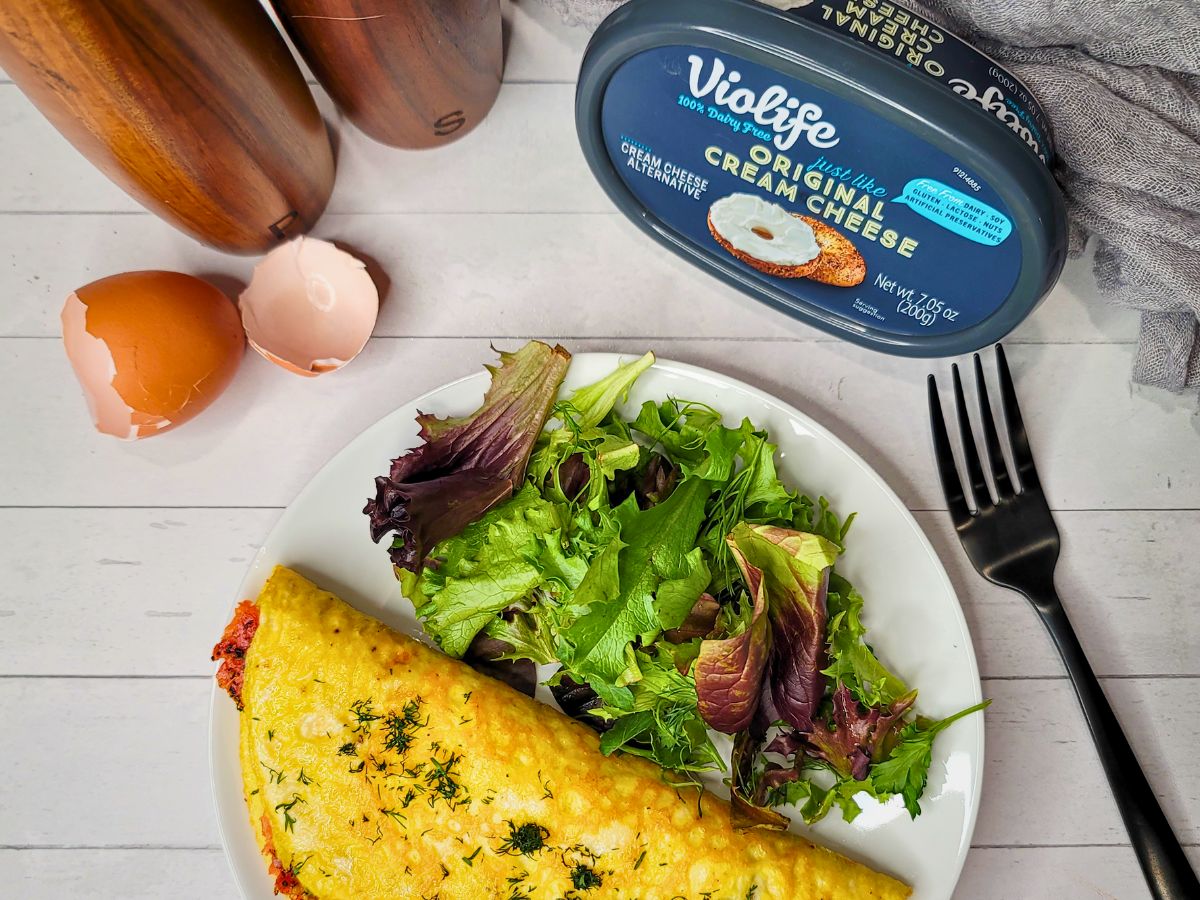 A plate with an omelette and mixed greens next to a fork, egg shells, pepper mills, and a container of Violife vegan cream cheese.