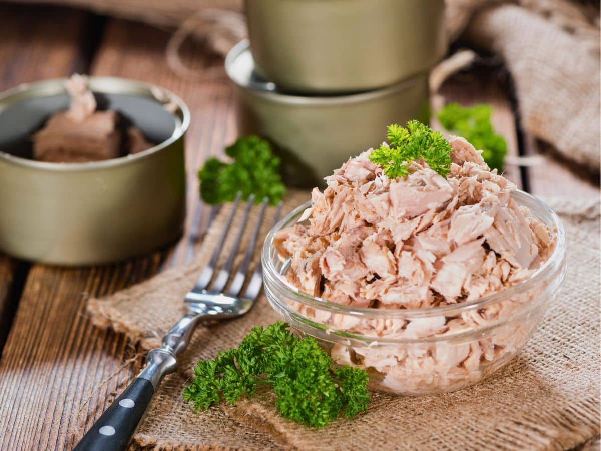 A clear glass bowl filled with flaked tuna, garnished with parsley, sits on a burlap cloth next to a fork and green ceramic bowls on a wooden table.