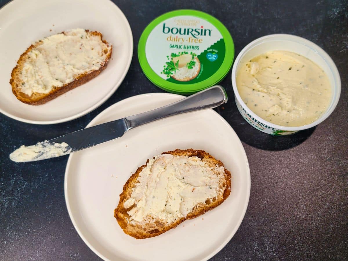 Two slices of bread with dairy-free Boursin garlic and herb spread on plates, with an open container and lid on a dark countertop. A knife with spread rests on one plate.