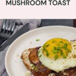 A slice of toast topped with sautéed mushrooms, feta cheese, and a sunny-side-up egg, garnished with chopped herbs, on a white plate.