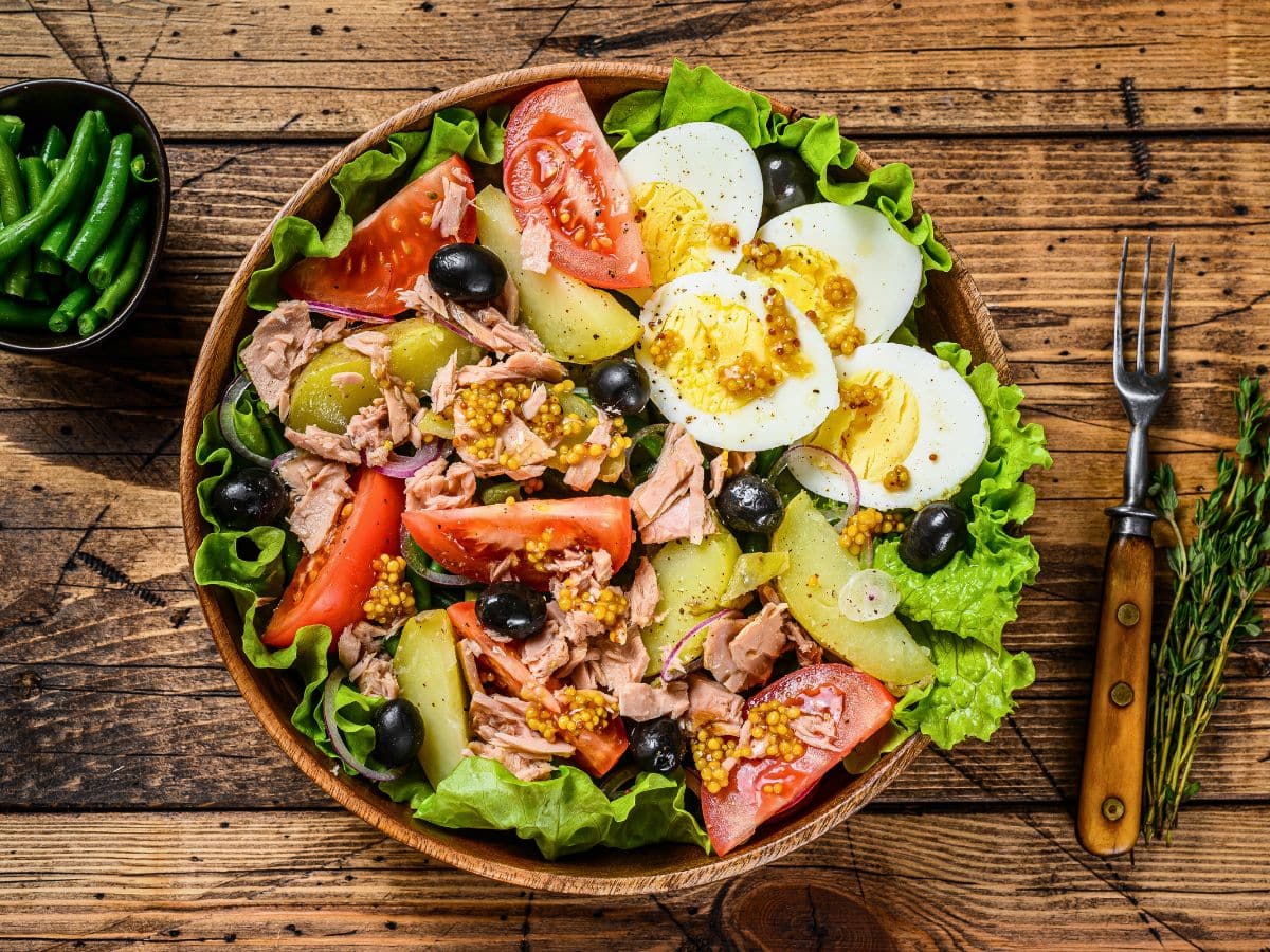 A bowl of salad with lettuce, tuna, tomato wedges, potato slices, hard-boiled egg slices, black olives, and dressing, on a wooden table with a fork and a small bowl of green beans.