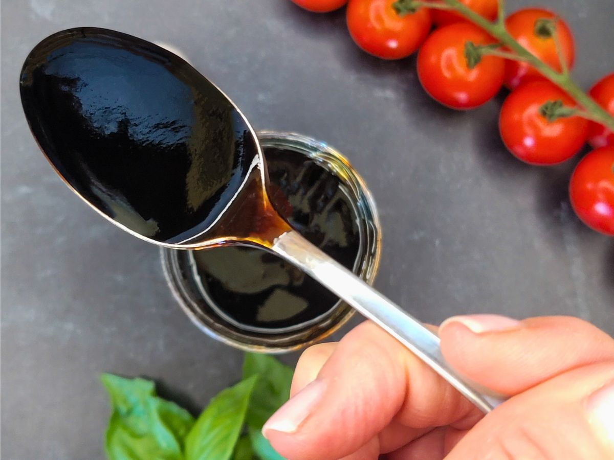 A hand holds a spoonful of thick, dark balsamic glaze over a jar, with vine tomatoes and basil leaves in the background on a dark surface.