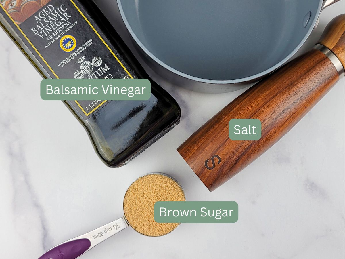 A bottle of balsamic vinegar, a salt grinder, a measuring cup of brown sugar, and a frying pan arranged on a white surface.