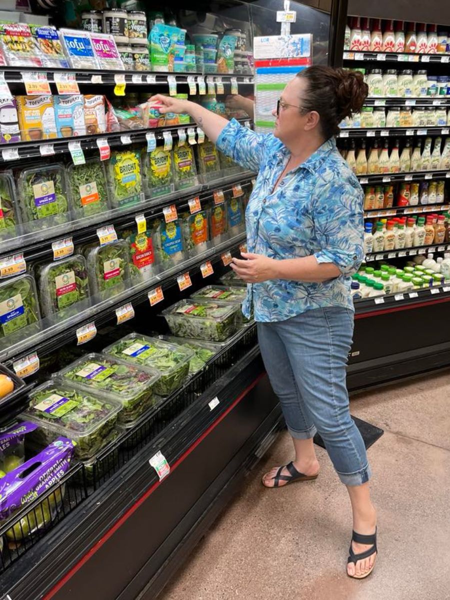 A woman in casual clothing selects a packaged salad from a refrigerated produce section in a grocery store.
