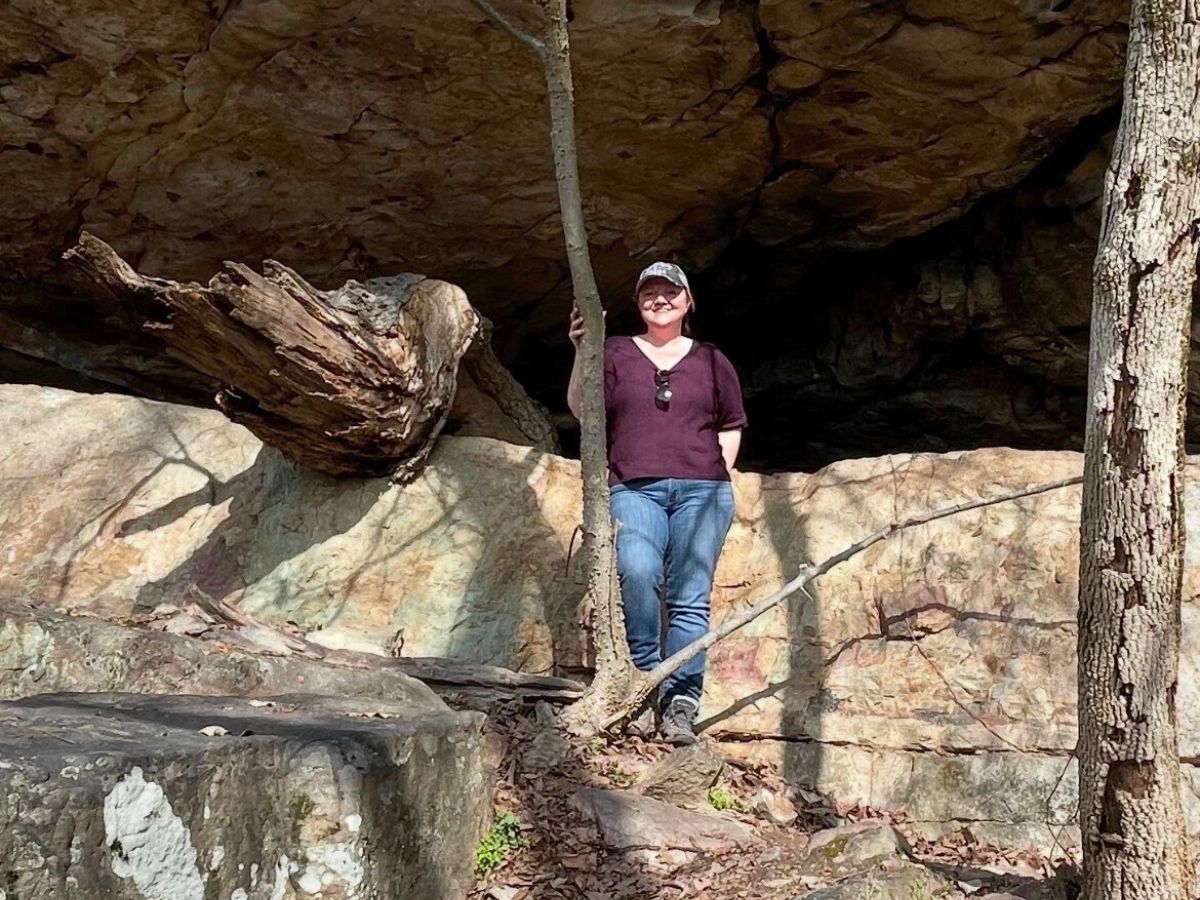 A person wearing a cap, sunglasses, and jeans stands outdoors near a rocky overhang and trees, with sunlight casting shadows.